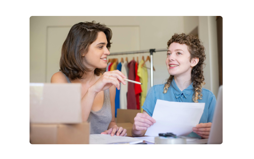 Dos mujeres planeando una tienda en línea para principiantes, con cajas de envío e inventario de ropa al fondo.