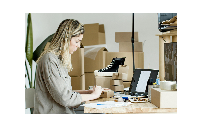Mujer administrando ventas de comercio electrónico, empaquetando pedidos con materiales de envío, cajas y una computadora portátil para el crecimiento del negocio en línea.